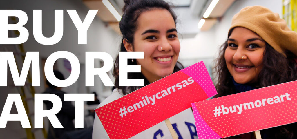 two students holding student art sale posters and posing. hashtags on the posters: #emilycarrsas #buymoreart